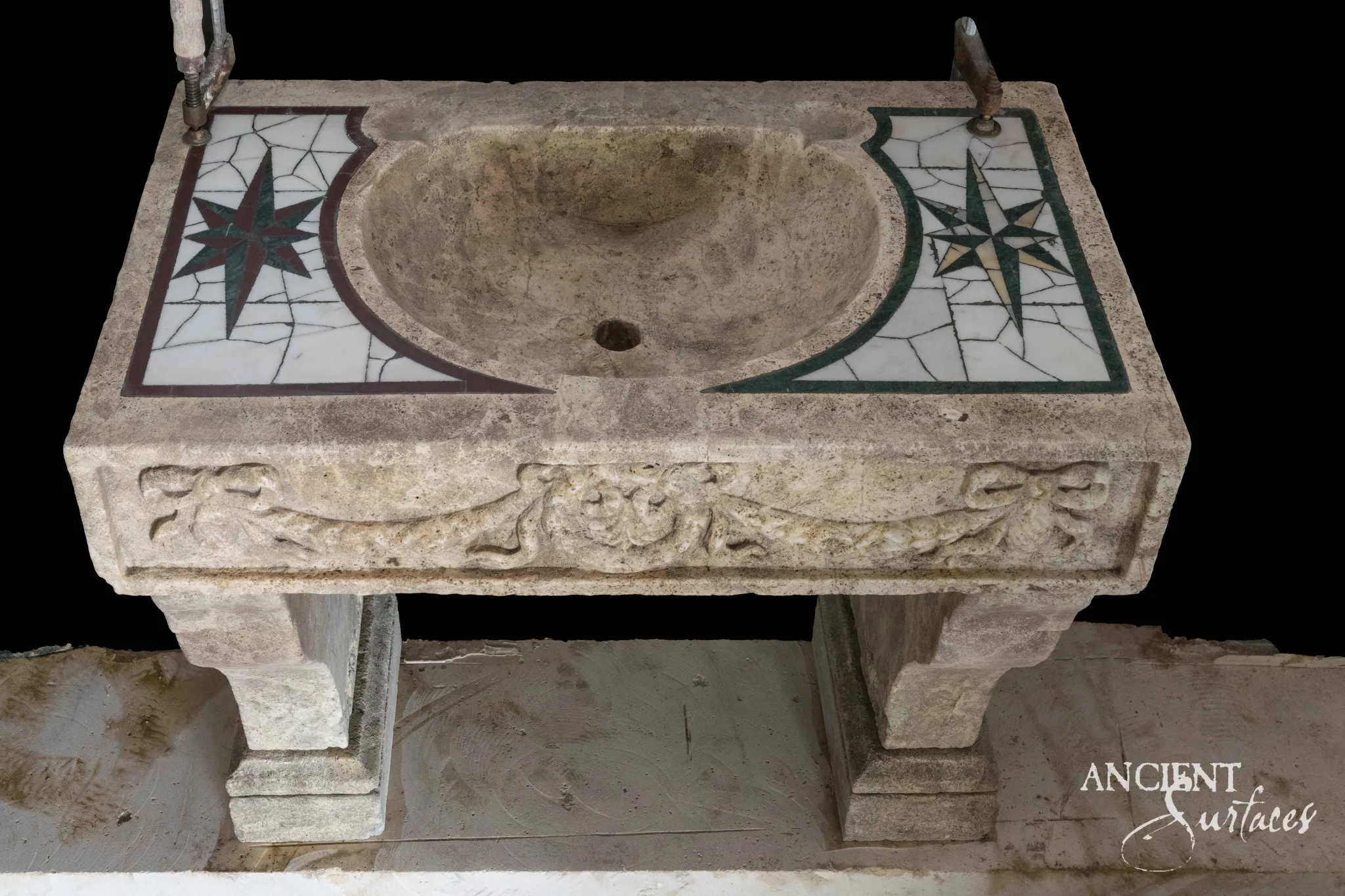 Reclaimed limestone sink seen from above, with a deep scooped bowl and drain between two inlaid compass star mosaic panels and a ribbon tied swag carved on the apron