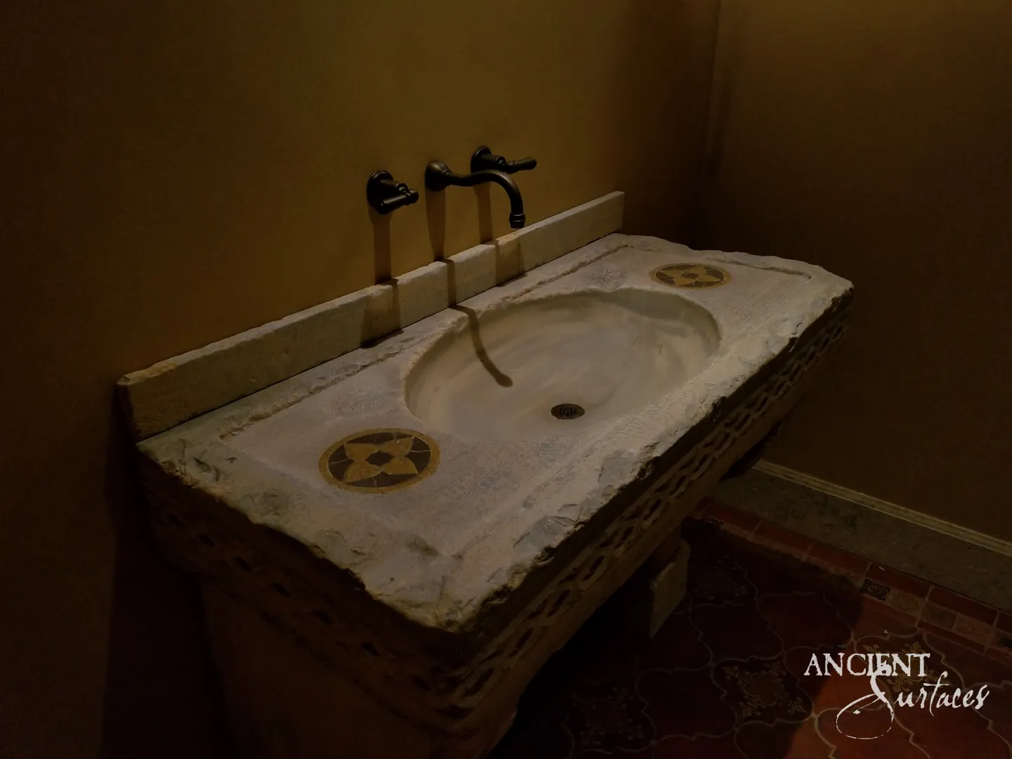 Reclaimed limestone slab sink in low light, with a wide oval bowl and drain, two inlaid four petal medallions, a lattice carved front edge and a low stone upstand