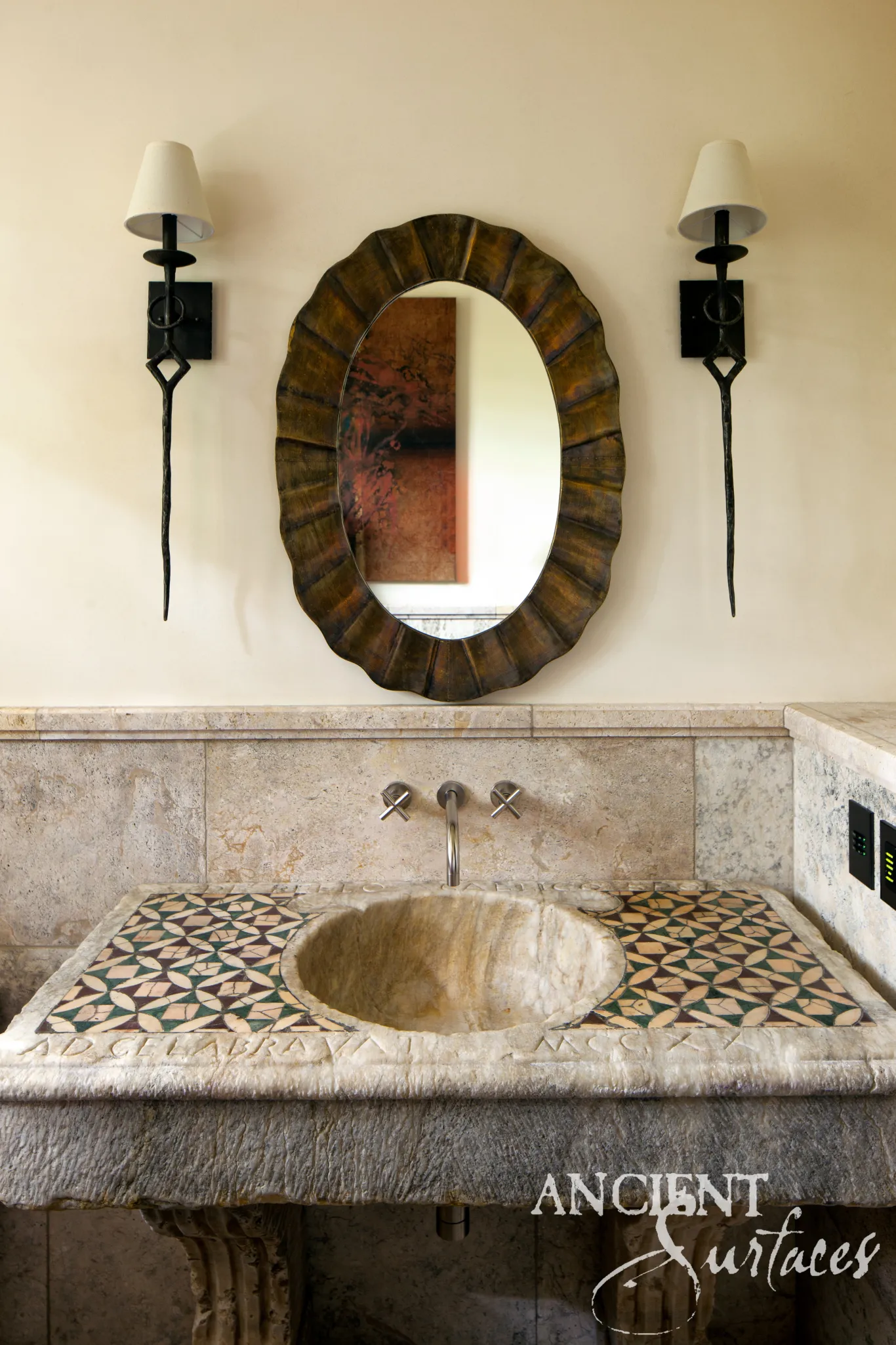 Reclaimed limestone sink installed under a wall mounted tap, with a geometric green and rose mosaic top, a deep round honey toned bowl, incised Roman lettering and a chisel dressed grey body