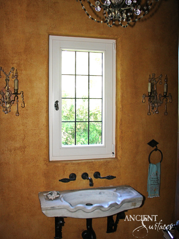 Slim reclaimed limestone wall sink with a scalloped front lip and shallow bowl, carried on dark iron brackets beneath a window in an ochre powder room